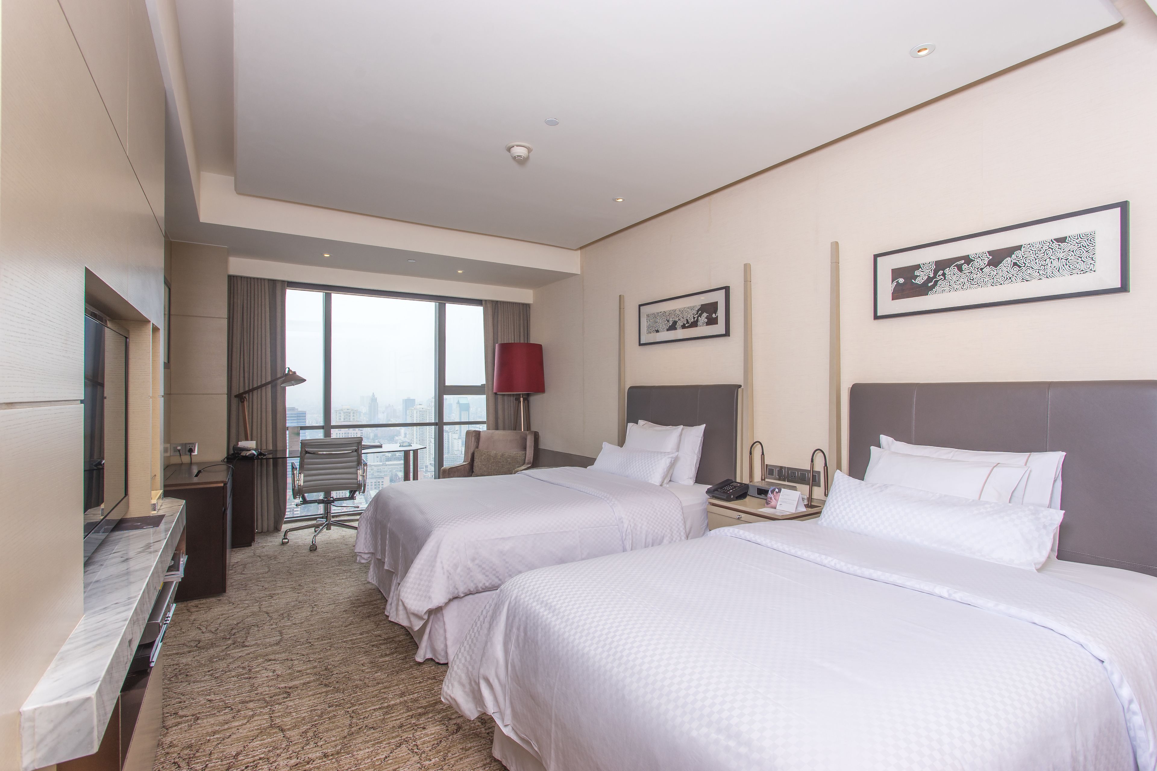 A hotel room with two beds, a desk, a chair, and a large window with a view of the city.