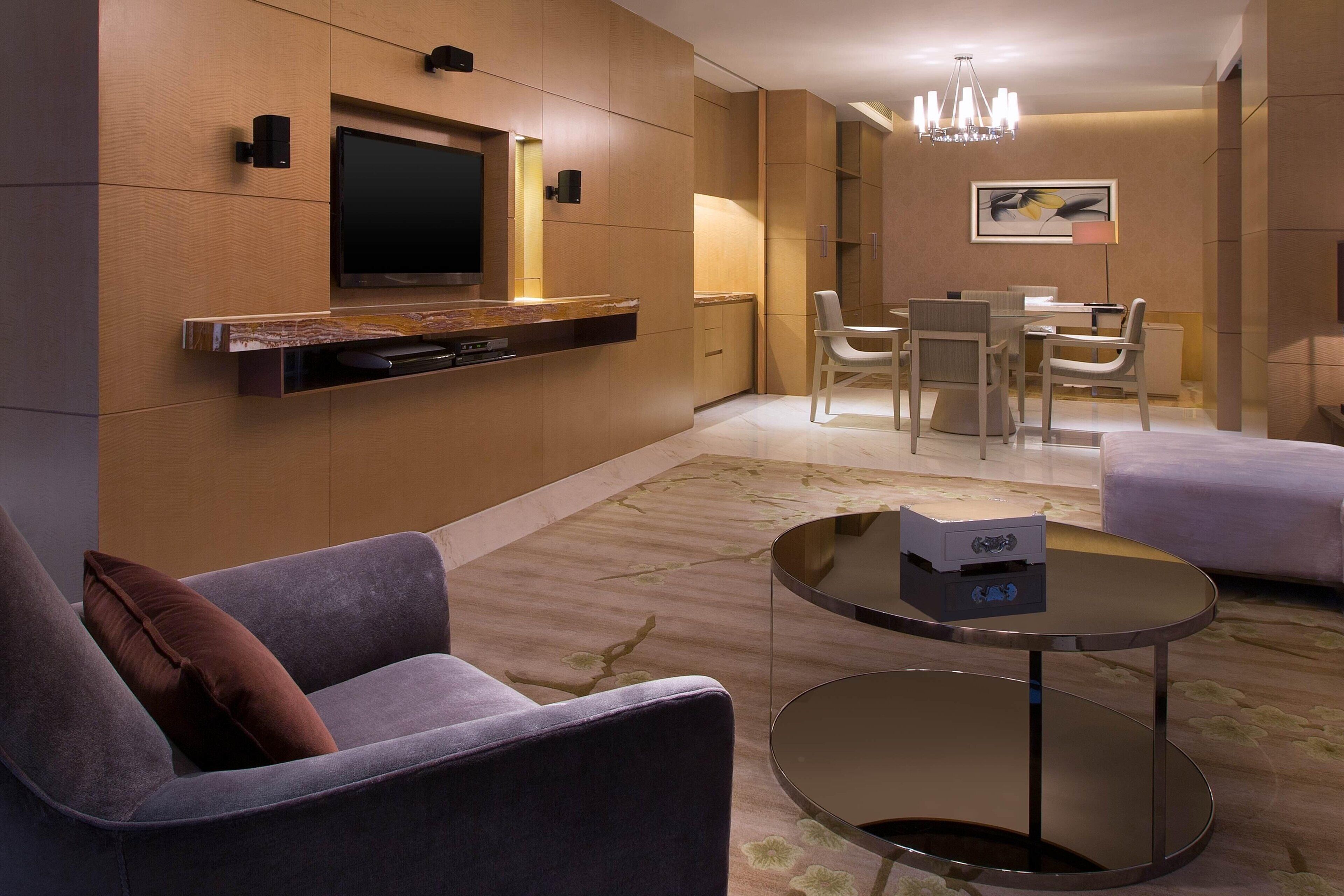 A modern living room with a flat-screen TV, a sofa, a coffee table, and a dining area with chairs and a table.