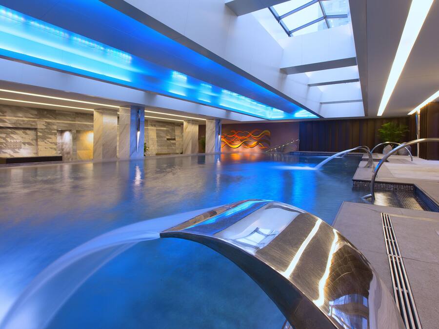 Un área interior de piscina moderna con techo de cristal, una bañera de hidromasaje y un espacio de descanso con una pared decorativa.