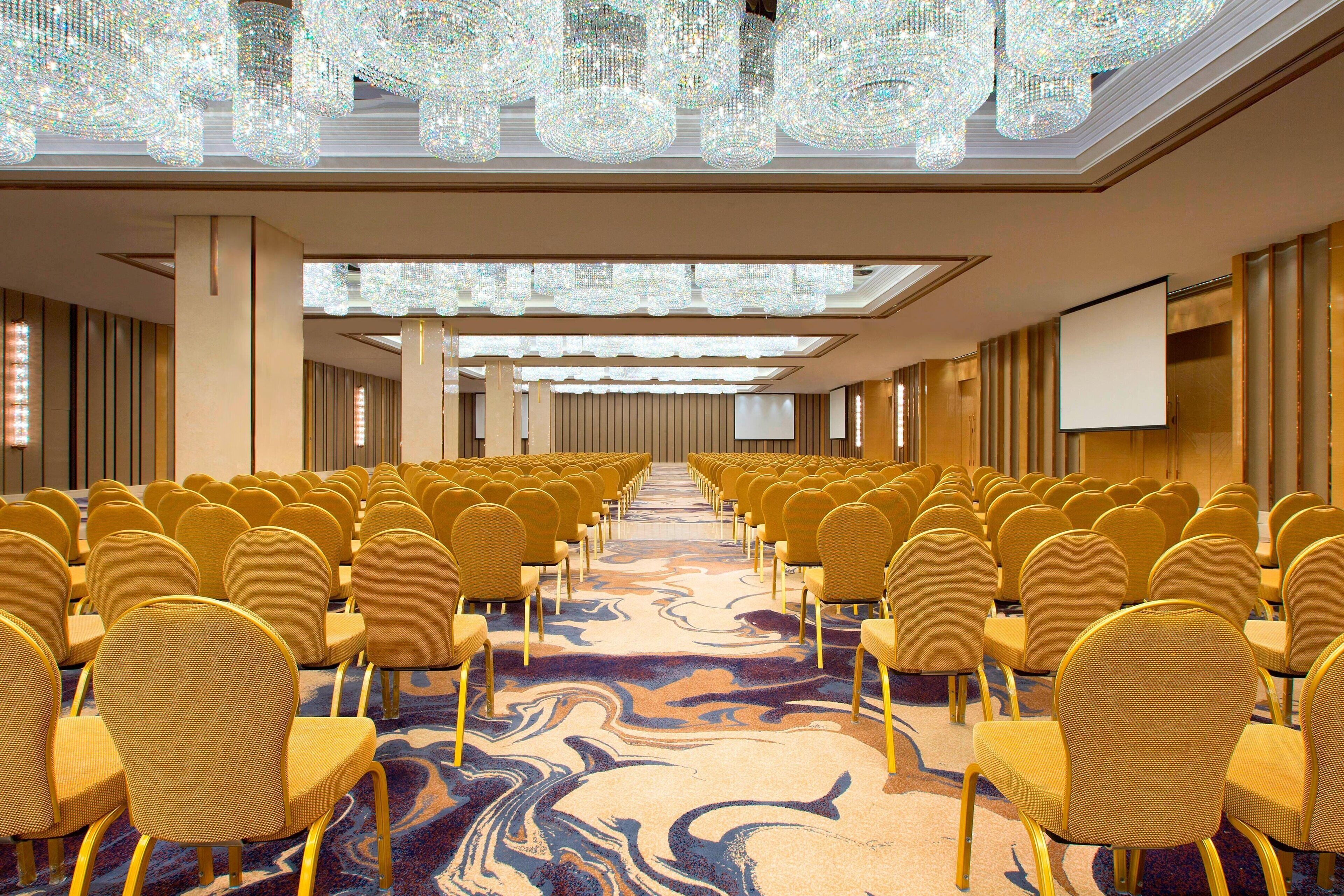 A conference room with rows of yellow chairs, a large screen, and chandeliers.