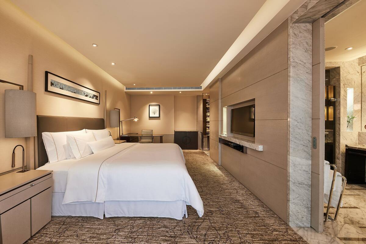 Une chambre d’hôtel moderne, dotée d’un grand lit, d’un bureau, d’une télévision et d’une salle de bain visible à travers une porte ouverte.