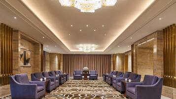 A conference room with a patterned carpet, rows of chairs, a chandelier, and a large painting on the wall.