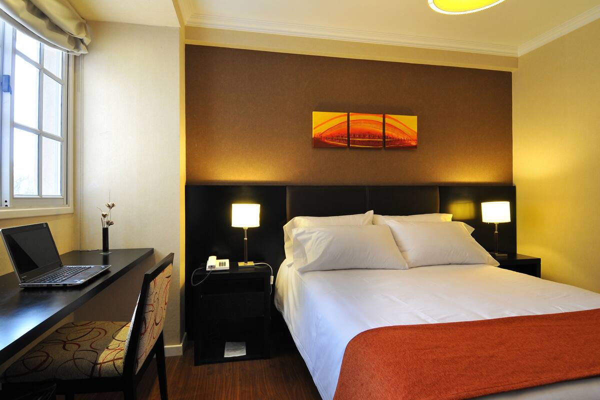 A hotel room with a bed, a desk with a laptop, two bedside lamps, and a wall art piece.