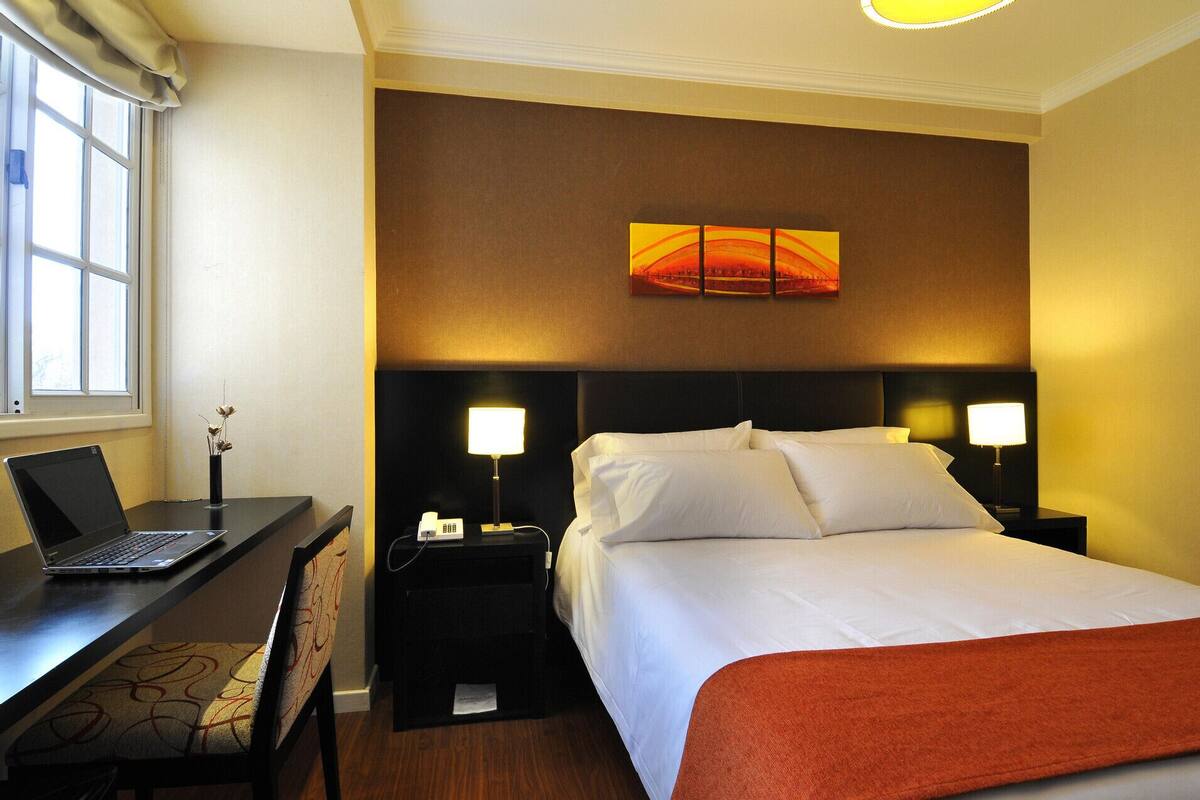 A hotel room with a bed, two bedside lamps, a desk with a laptop, and a painting on the wall.