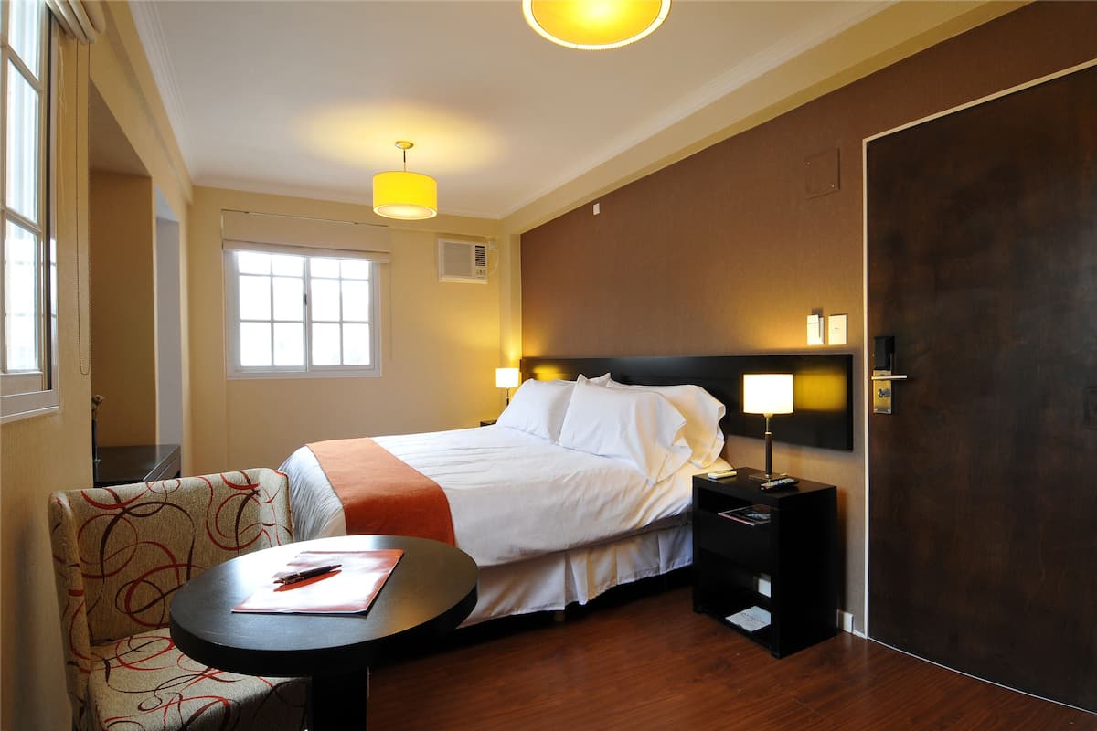 A hotel room with a bed, bedside table, lamp, chair, and a window.