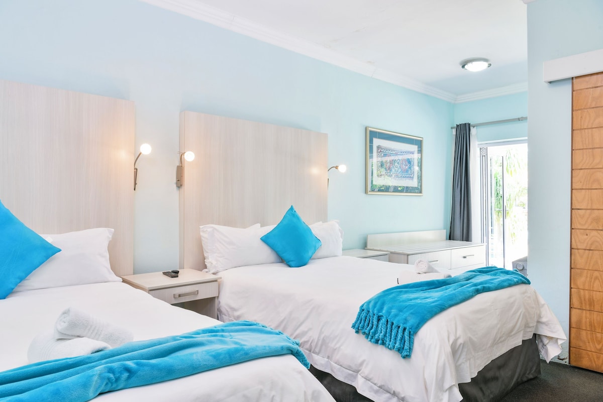 A hotel room with two beds, each with white bedding and blue pillows. There is a wooden wardrobe, a nightstand with a lamp, and a framed picture on the wall.