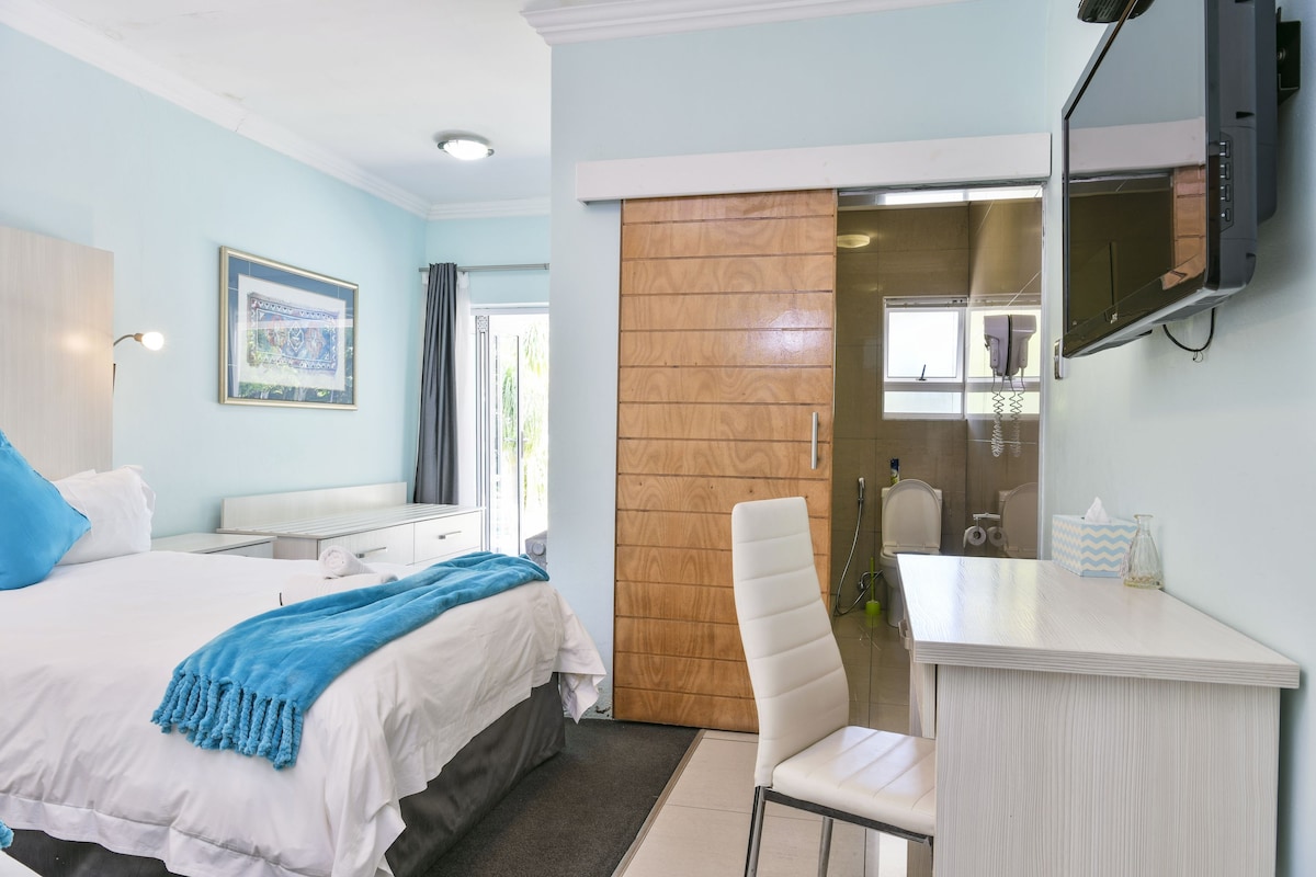 A hotel room with a bed, a desk, a mirror, and a bathroom visible through a sliding door.
