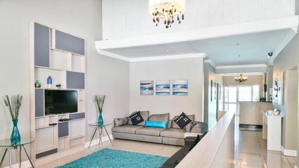 A modern living room with a flat-screen TV, a grey sofa with blue cushions, a teal rug, and a staircase leading to an upper level. - Al-Beit Lodge (Gqeberha (Port Elizabeth))