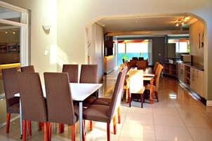 A dining area with a long table and chairs, a kitchen in the background, and a view of the outdoors.