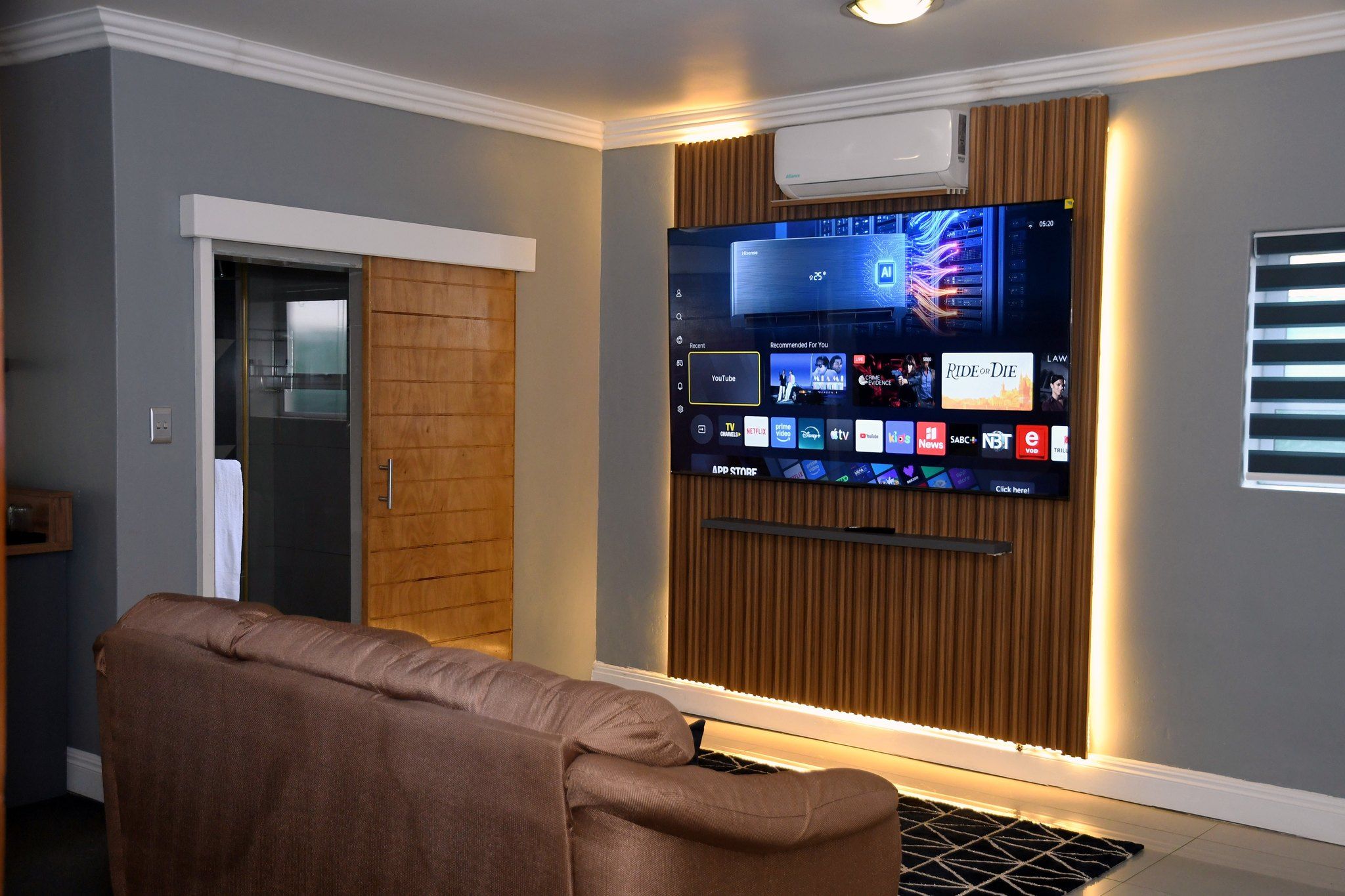 A modern living room with a flat-screen TV mounted on a wall, a brown sofa, and a wooden panel design.
