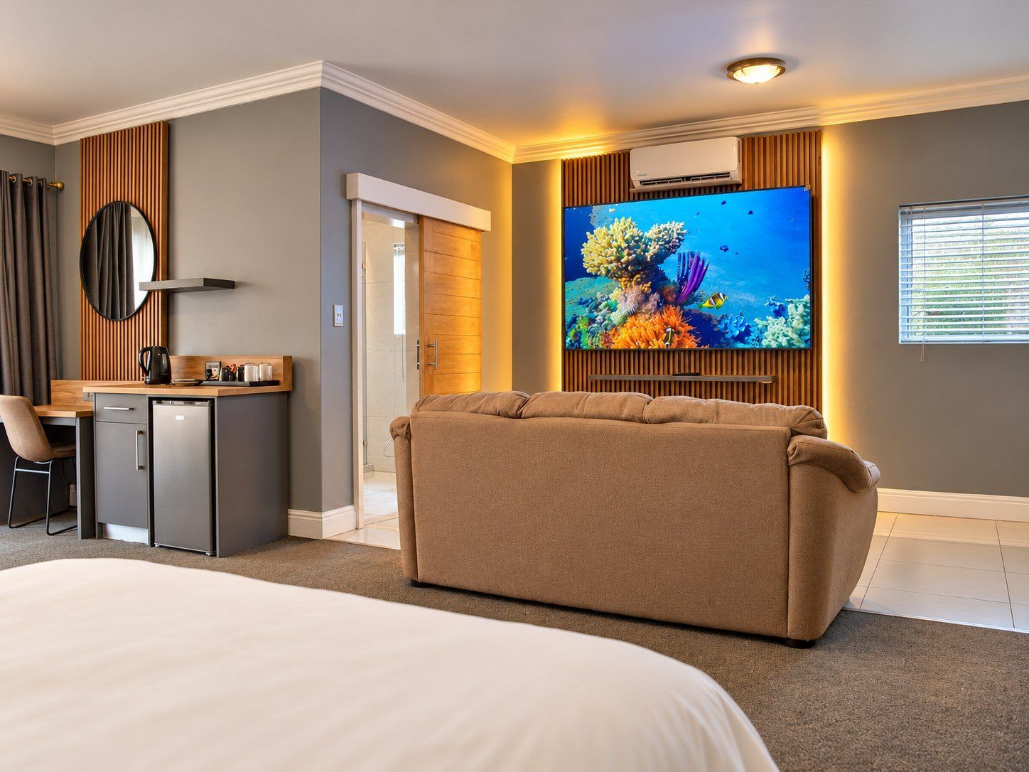 A hotel room with a bed, a seating area, a desk, a television, and an air conditioning unit.