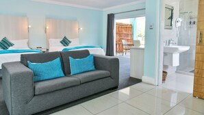 A modern hotel room with a grey sofa, two beds with blue and white bedding, a bathroom with a glass shower, and a view of a wooden fence outside. - Al-Beit Lodge (Gqeberha (Port Elizabeth))