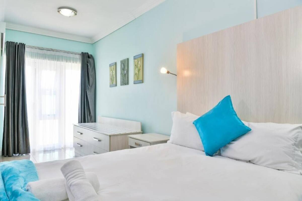 Ein Hotelzimmer mit Bett, Schreibtisch und Nachttisch. Am Fenster hängen Vorhänge und an der Wand ist eine Leuchte befestigt.
