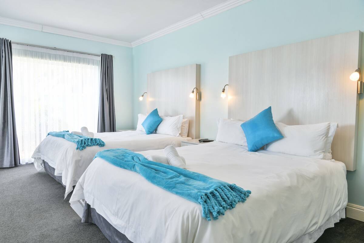 Ein Hotelzimmer mit zwei Betten, hellblauen Wänden und einem großen Fenster mit Vorhängen.