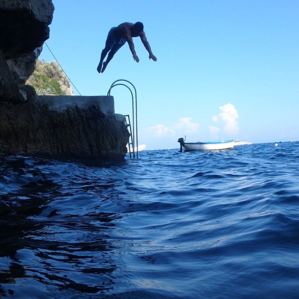 A person diving from a rocky platform into the ocean, with boats in the background.
