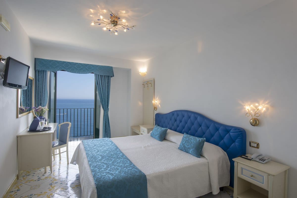 Une chambre d’hôtel avec un grand lit, un bureau avec une chaise, une télévision et une vue sur la mer depuis un balcon.