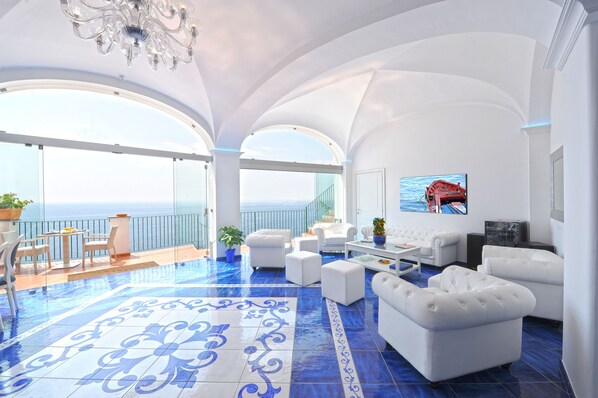 A spacious living area with a large balcony overlooking the sea, featuring white furniture and a tiled floor.