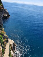 Un sendero costero con una escalera de piedra que desciende al mar, rodeado de vegetación y con vista despejada del océano.