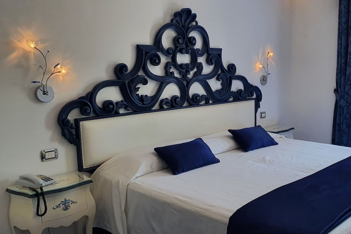 Un lit bien fait, recouvert d’une couvre-lit blanche et bleue, agrémenté d’une tête de lit décorative, avec un téléphone sur la table de chevet et une vue sur un balcon extérieur équipé de chaises.