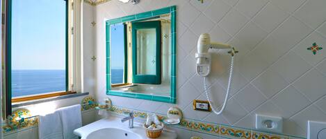 A bathroom with a large sink, a mirror, a hairdryer, and a window with a view of the sea.