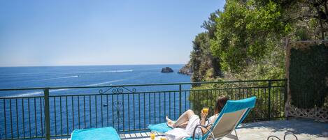 A balcony with a view of the sea, a table with food and drinks, and two lounge chairs.