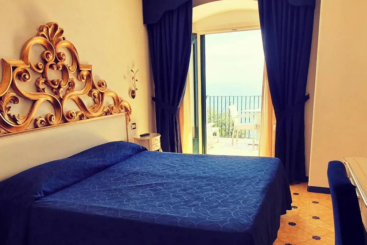 Une chambre d’hôtel avec un grand lit, une tête de lit décorative, une petite table de chevet et une vue sur la mer depuis un balcon.