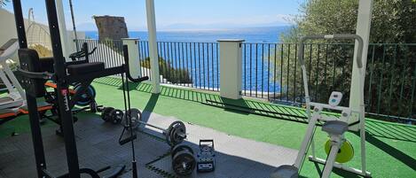 Un espace de remise en forme extérieur abrité, équipé de matériel d’exercice, offrant une vue sur la mer et doté d’un sol en gazon synthétique vert.