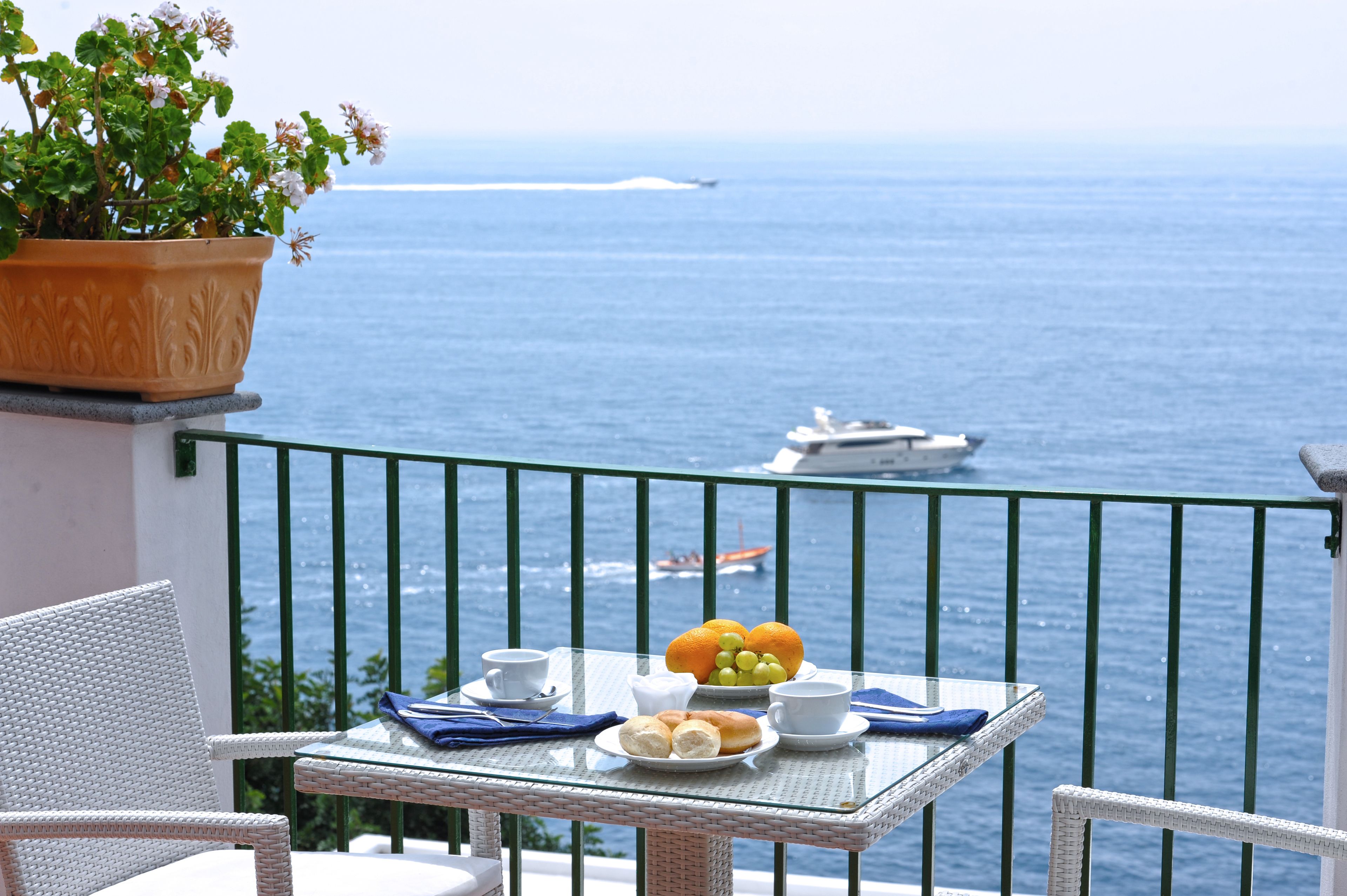 Un balcón con una mesa lista para el desayuno, con vista al mar y un barco a lo lejos.