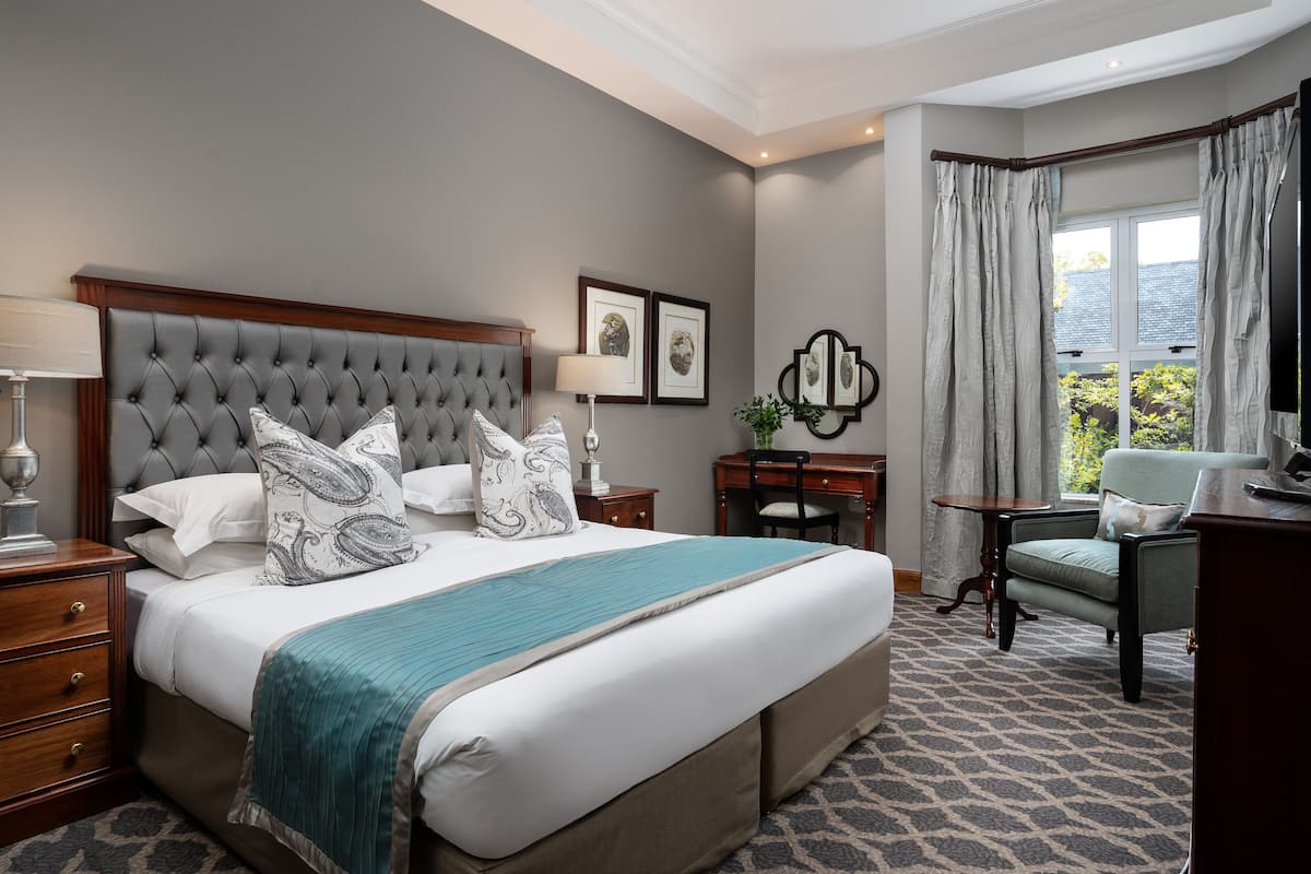 Een netjes ingerichte slaapkamer met een gestoffeerd hoofdbord, een groot bed met meerdere kussens, een nachtkastje met een lamp, een stoel, en uitzicht op groen door een raam met gordijnen.