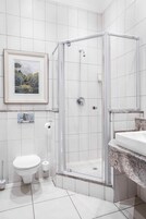 A bathroom with white tiles, a framed picture on the wall, a toilet, a shower with a glass door, and a sink with a granite countertop.