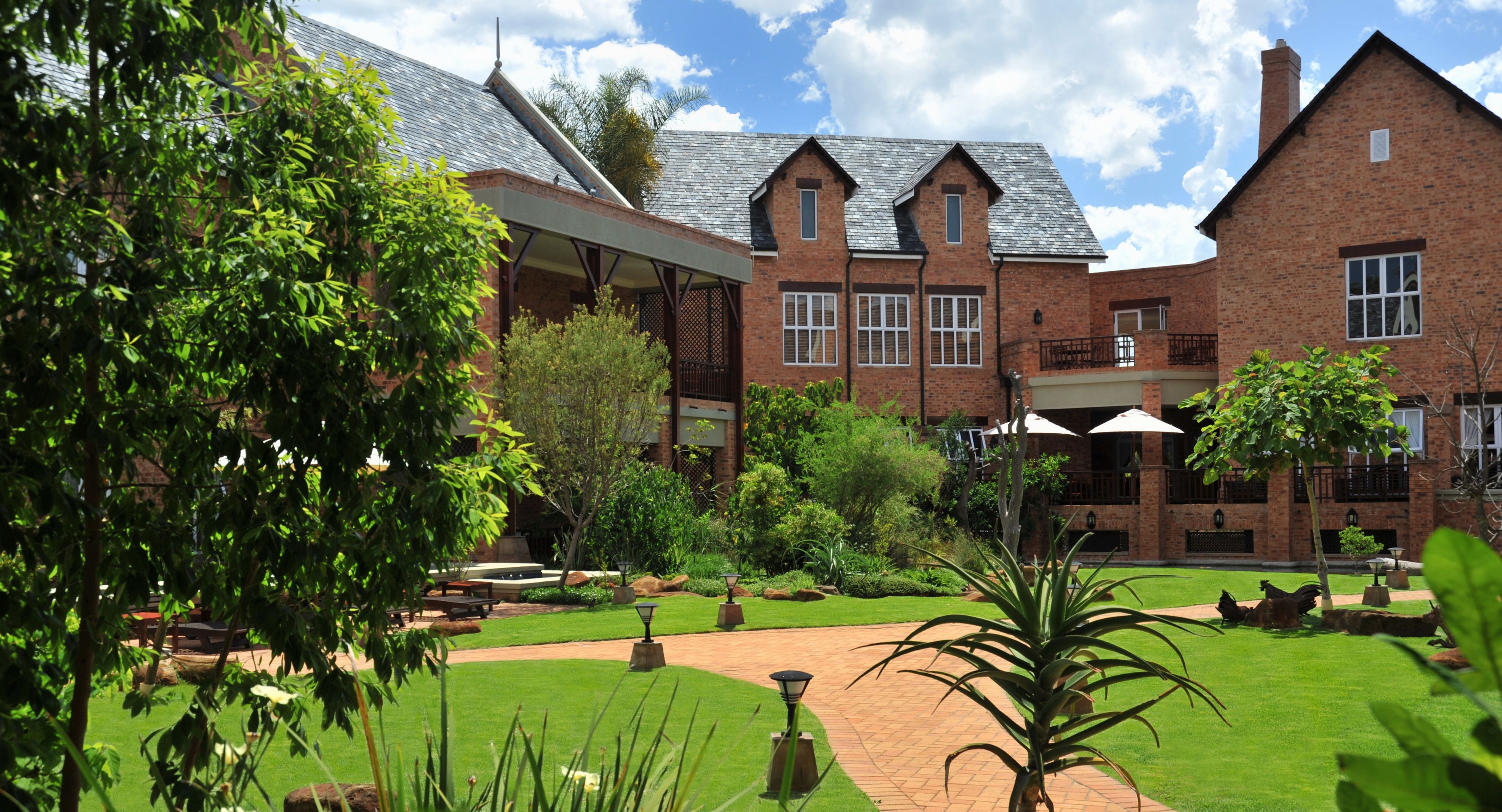 A large brick building with multiple windows, a covered patio area, and a well-maintained garden with pathways and seating.