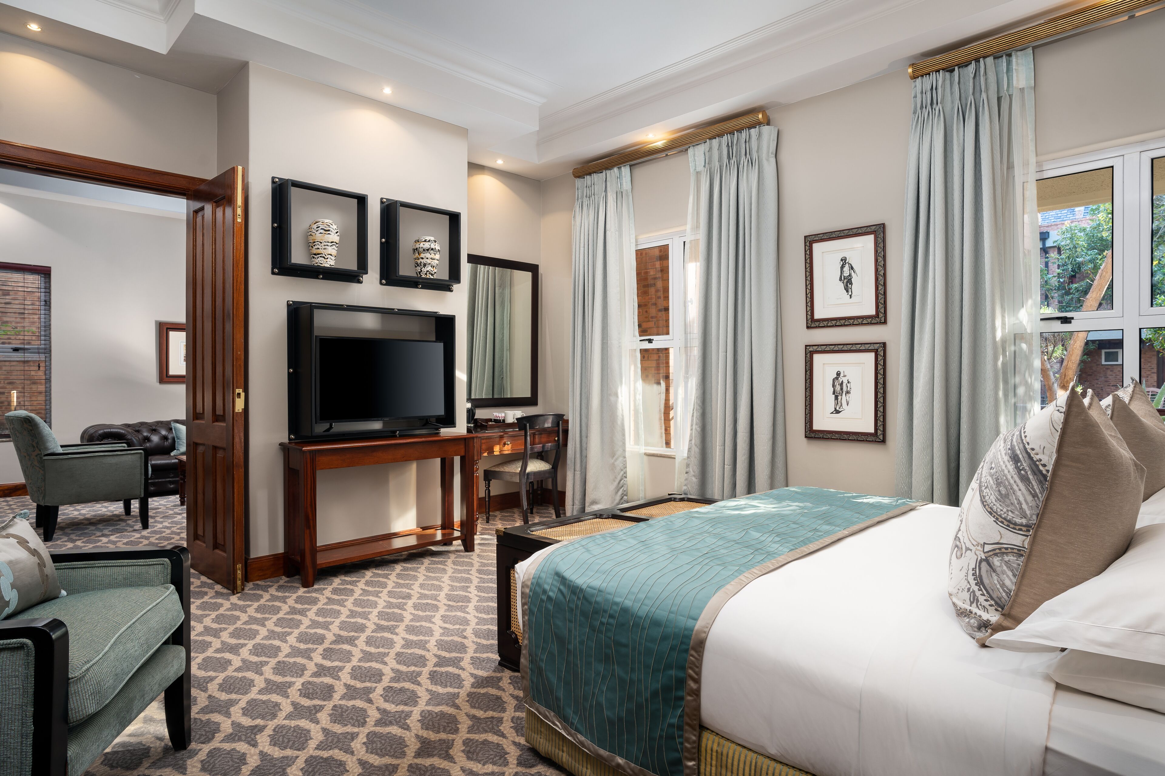 A hotel room with a large bed, a desk with a TV, a seating area with chairs, and framed pictures on the walls.