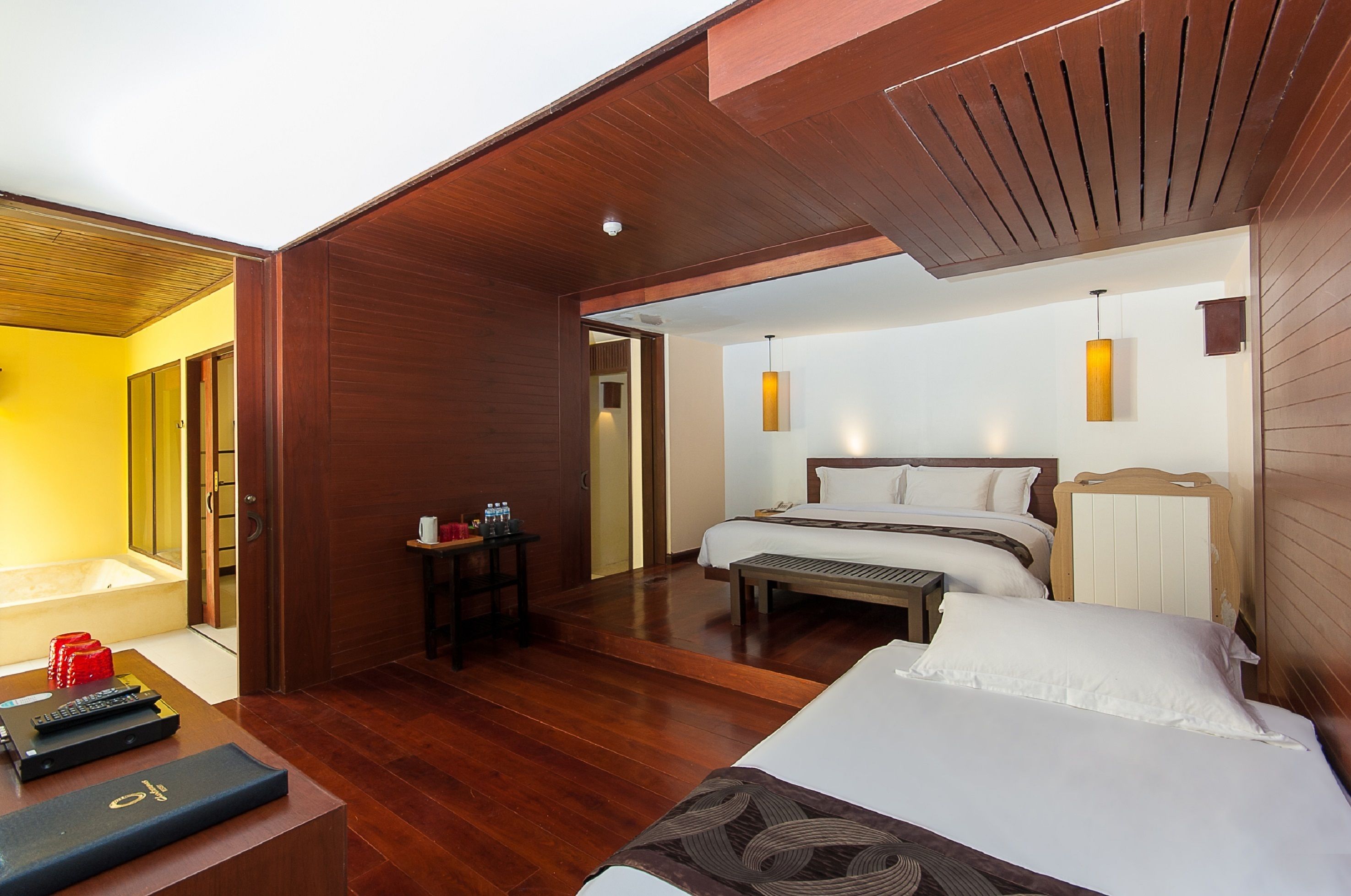 Quarto de hotel com teto de madeira, cama com roupa de cama branca, mesa de cabeceira com abajur e uma pequena mesa com garrafa e copos.