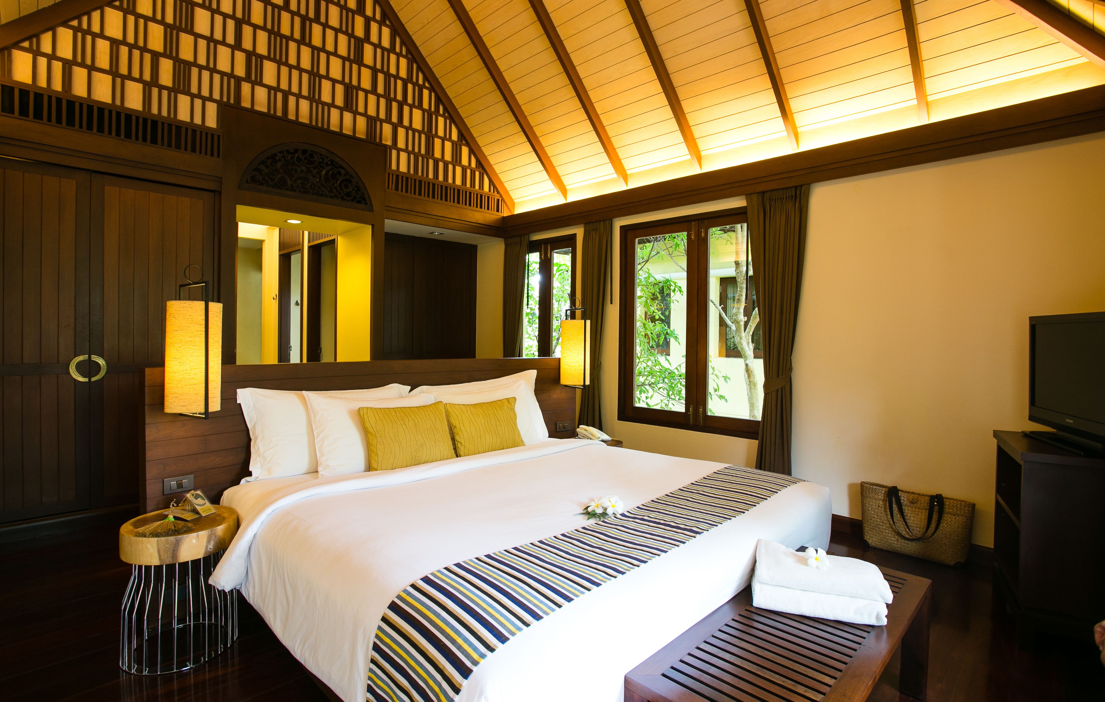 A hotel room with a large bed, a wooden headboard, a bedside table with a lamp, a TV, and a window with curtains.