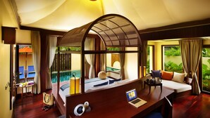 Habitación de hotel con cama, escritorio con computadora portátil, una mesita de noche con lámpara y vistas a una piscina y follaje.
