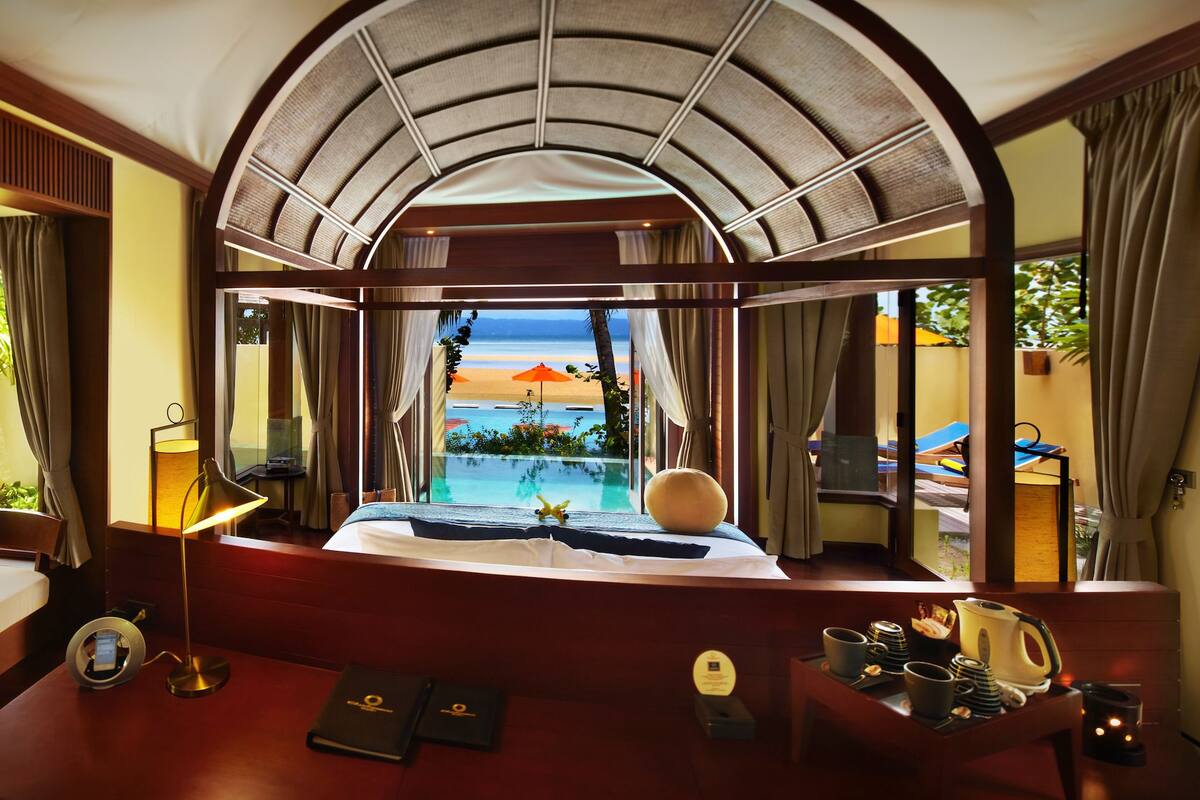 A luxurious room with a large window overlooking a pool and beach, a sofa, a desk with a lamp, and a tray with coffee and pastries.