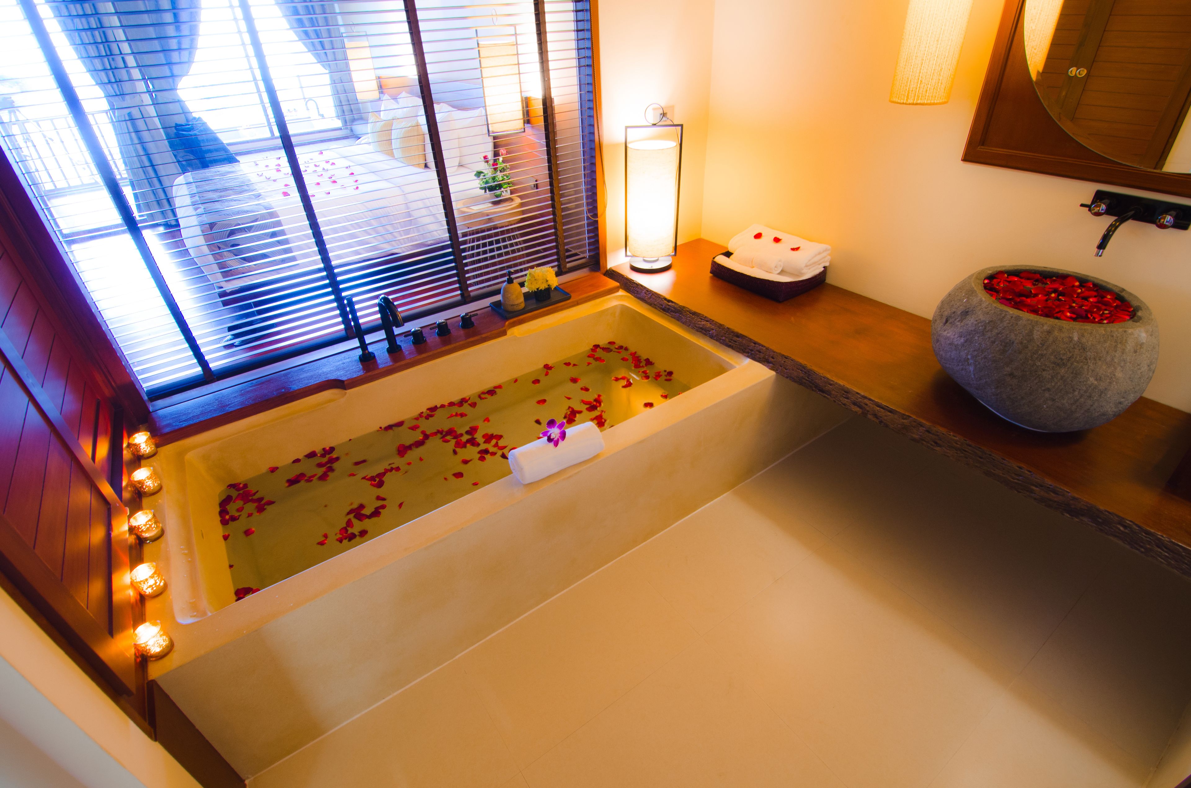 Une salle de bain avec une baignoire remplie de pétales de roses, un lavabo en pierre orné de pétales rouges et une fenêtre avec des stores.