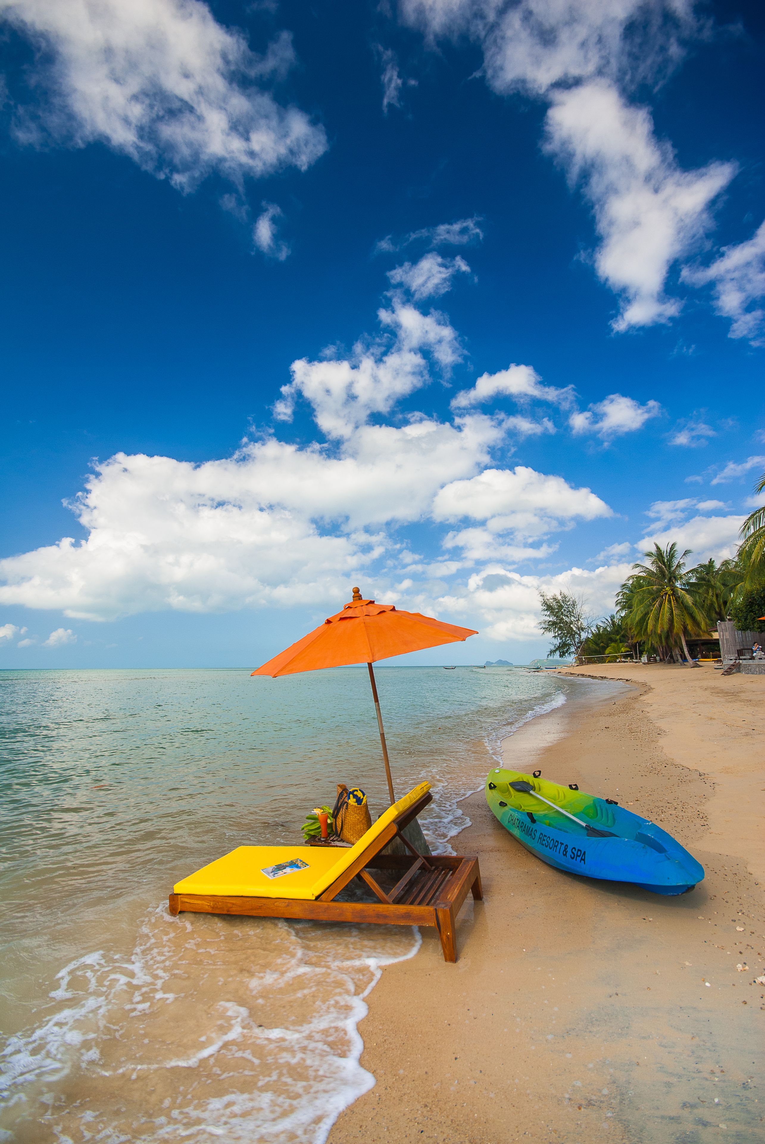 Una playa con una silla de playa amarilla y un paraguas naranja, un kayak azul y palmeras.