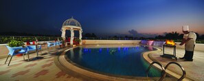 A pool area with a gazebo, outdoor seating, and a chef preparing food.