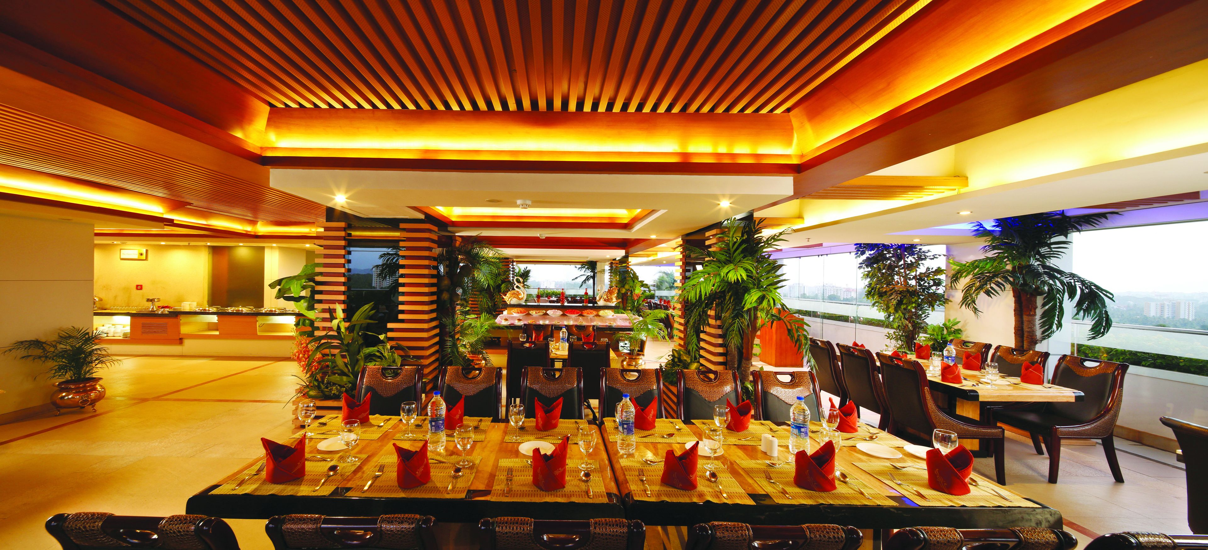 A restaurant with a long dining table set for a meal, surrounded by chairs, and decorated with plants and red napkins.