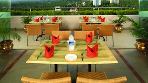 A dining area with tables set for four, each with red napkins, a bottle of Aquafina water, and a white plate. There are wicker chairs and potted plants. - Windsor Rajadhani (Thiruvananthapuram)
