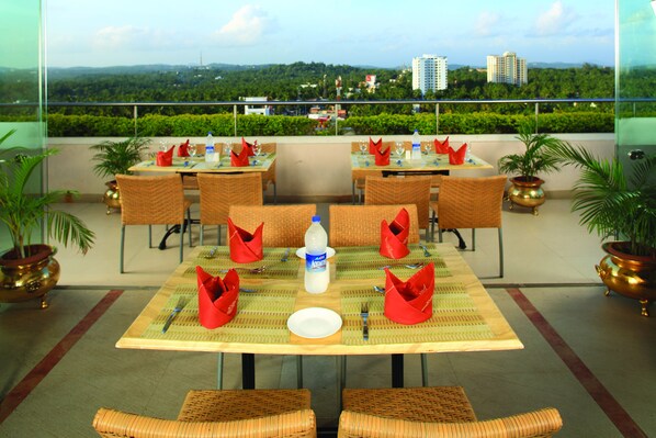 A dining area with tables set for four, each with red napkins, a bottle of Aquafina water, and a white plate. There are wicker chairs and potted plants.