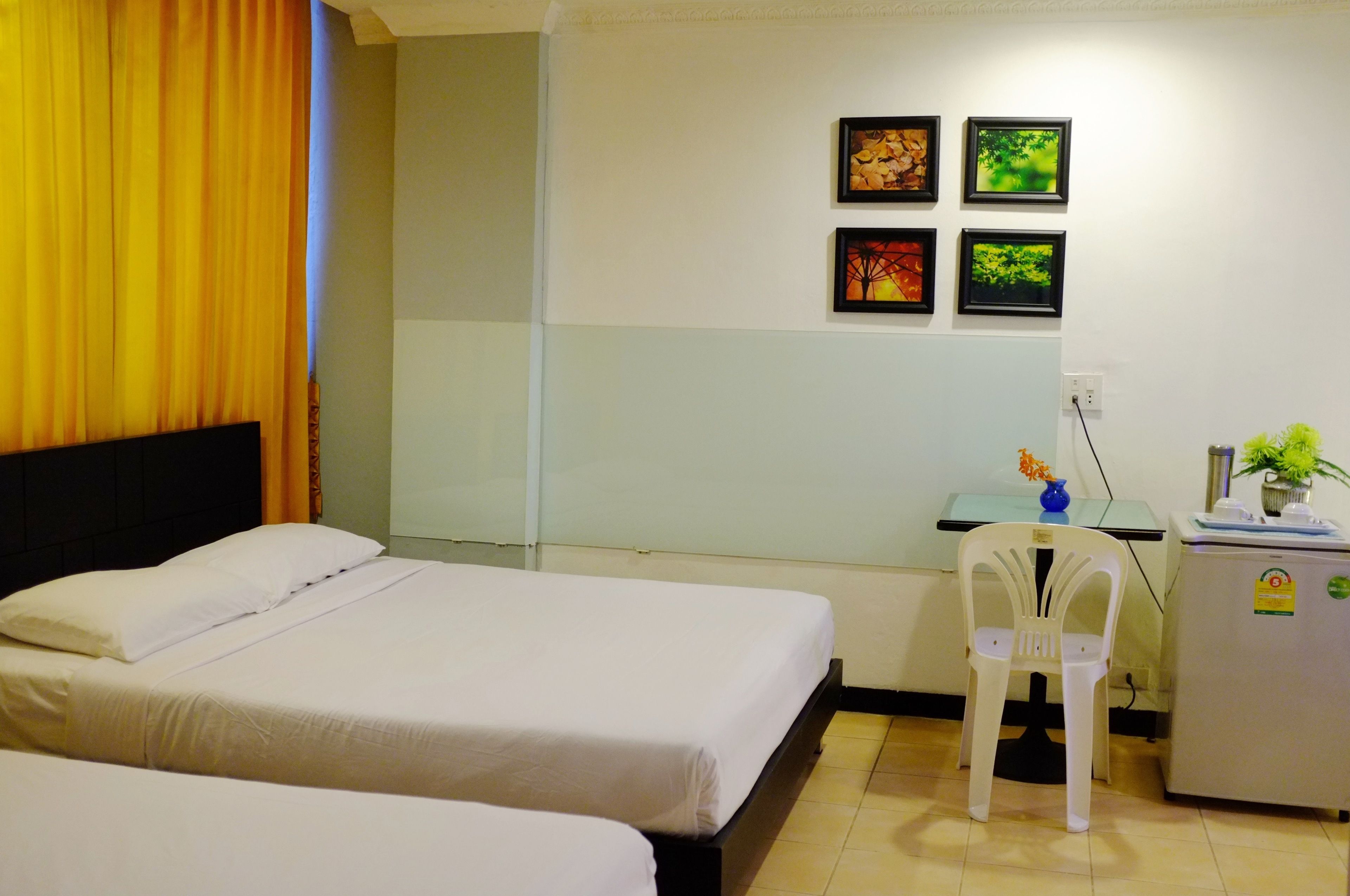 Habitación de hotel con dos camas, un escritorio con silla, un refrigerador pequeño y cuatro cuadros en la pared.