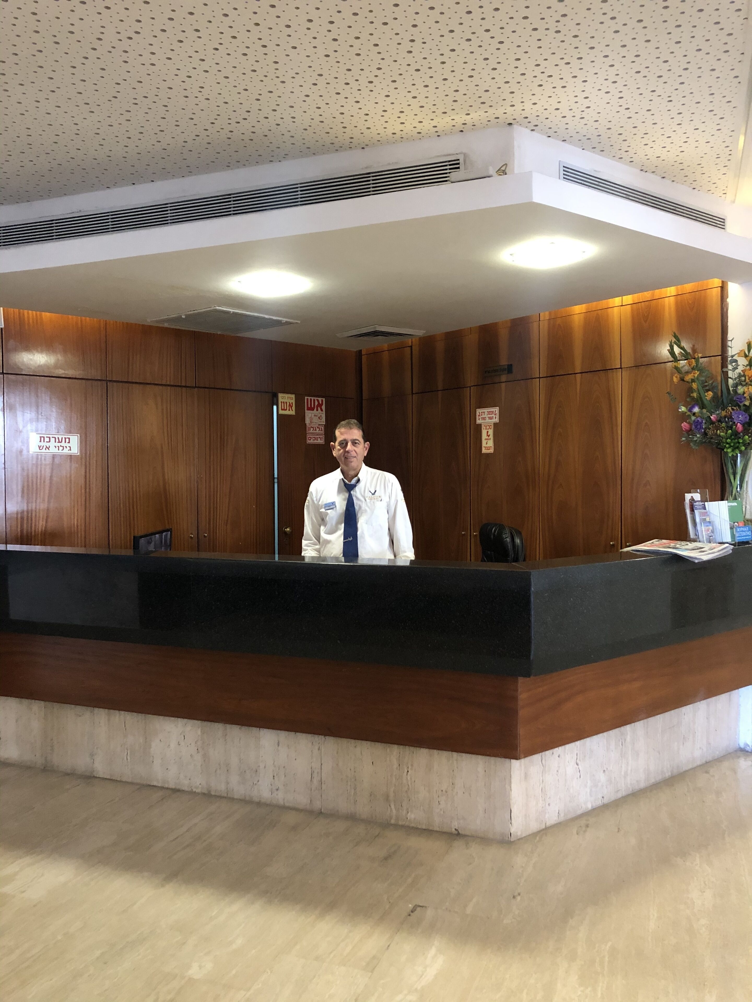 A reception desk with a man standing behind it, wooden paneling on the walls, and a ceiling with a perforated pattern.