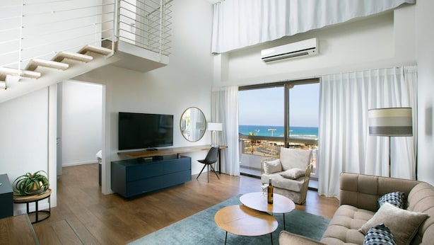 A modern living room with a TV, sofa, and coffee table, a staircase, and a large window with a view of the sea. - Marine Heights Suites (Herzliya)
