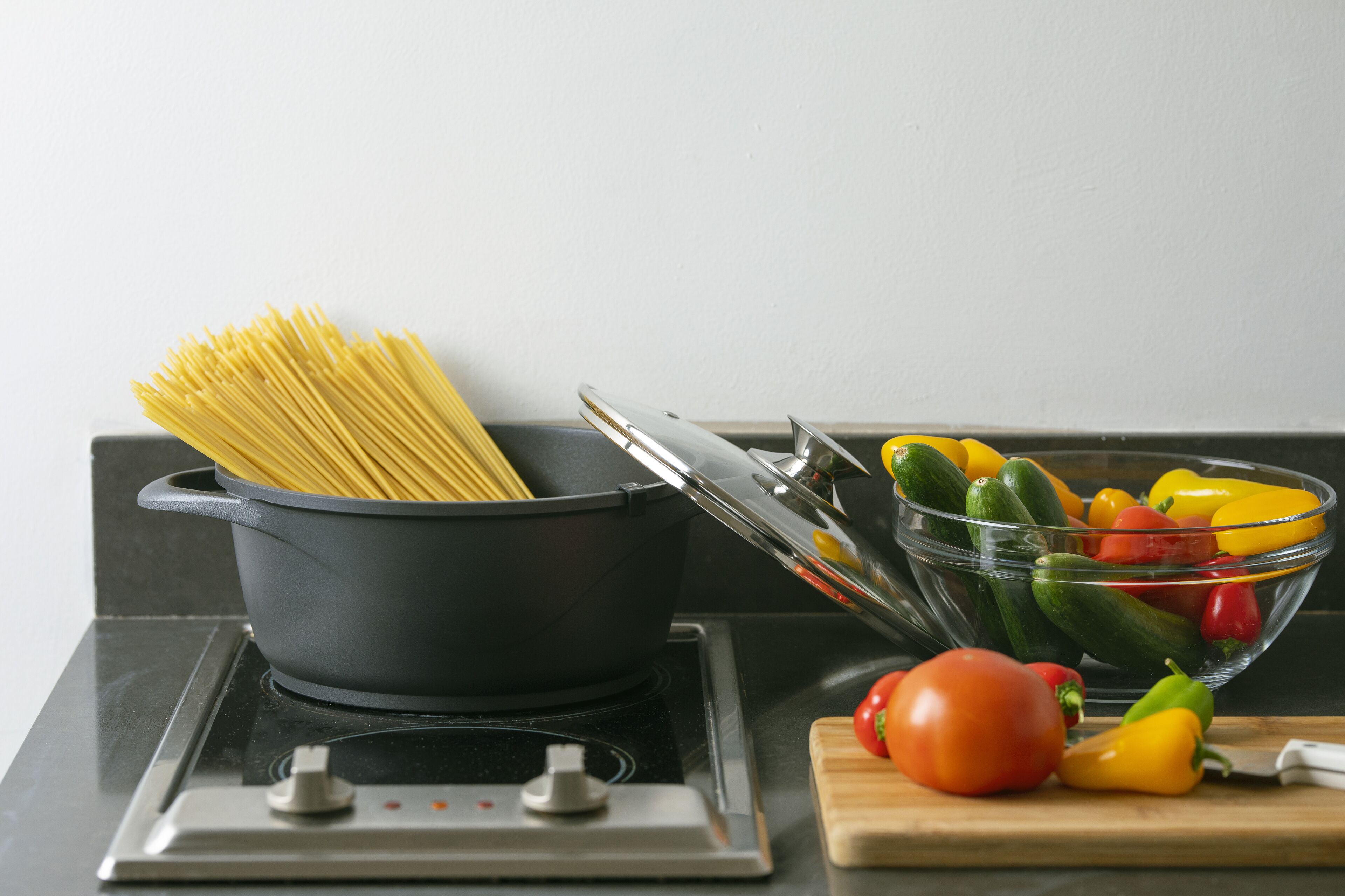 A kitchen with a pot of spaghetti, a bowl of vegetables, and a tomato on a cutting board.