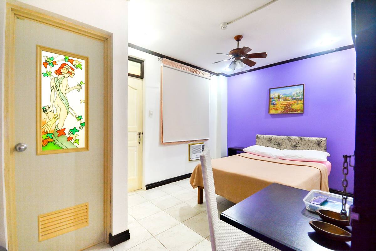 Una habitación con cama, una mesa con balanza, una silla, una pizarra y un ventilador de techo.