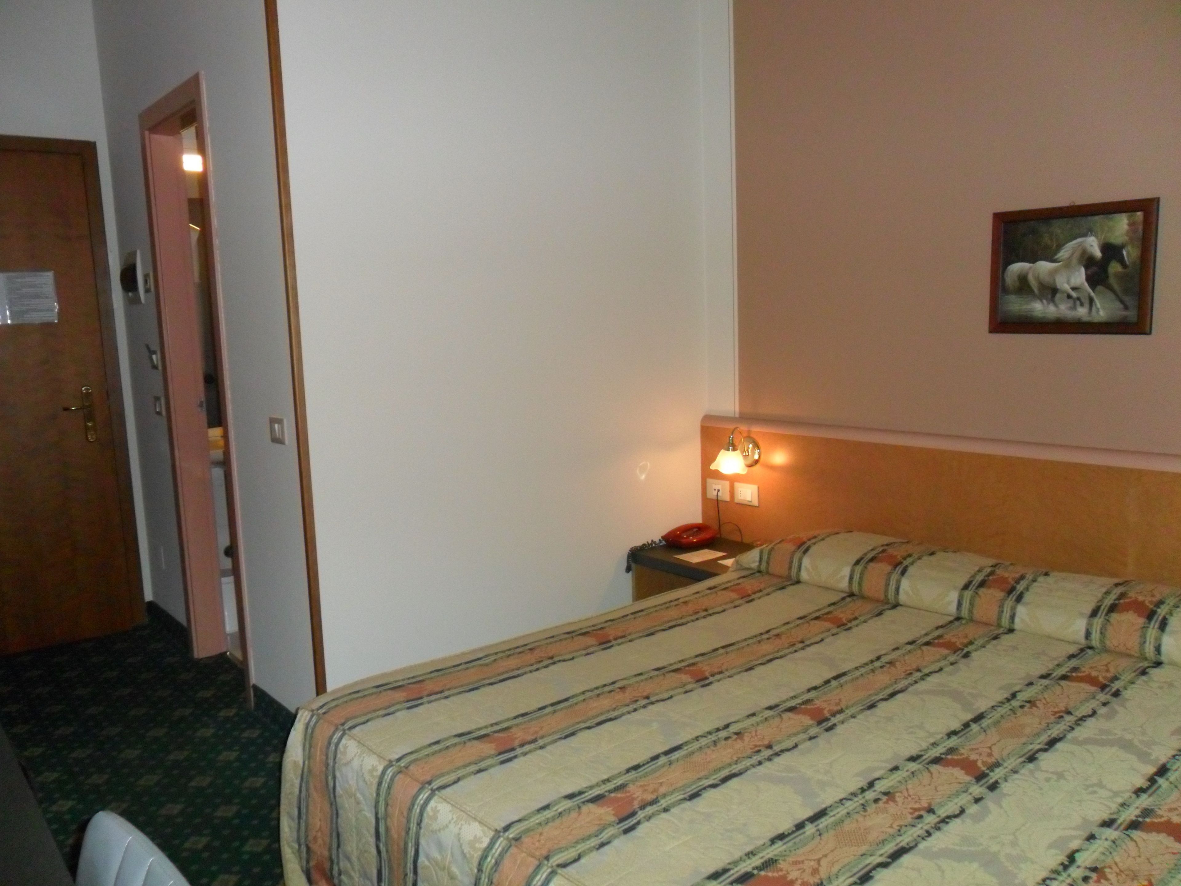 Ein Hotelzimmer mit einem Bett, einem Nachttisch, einer Lampe, einem Bild und einer Tür.