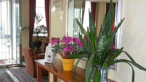 A reception area with a wooden console table, a potted plant, a mirror, and a red patterned rug. - Hotel Riviera (Segrate)