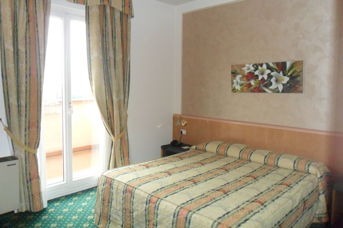 A hotel room with a bed, a nightstand, a painting of flowers, and a sliding glass door with curtains.